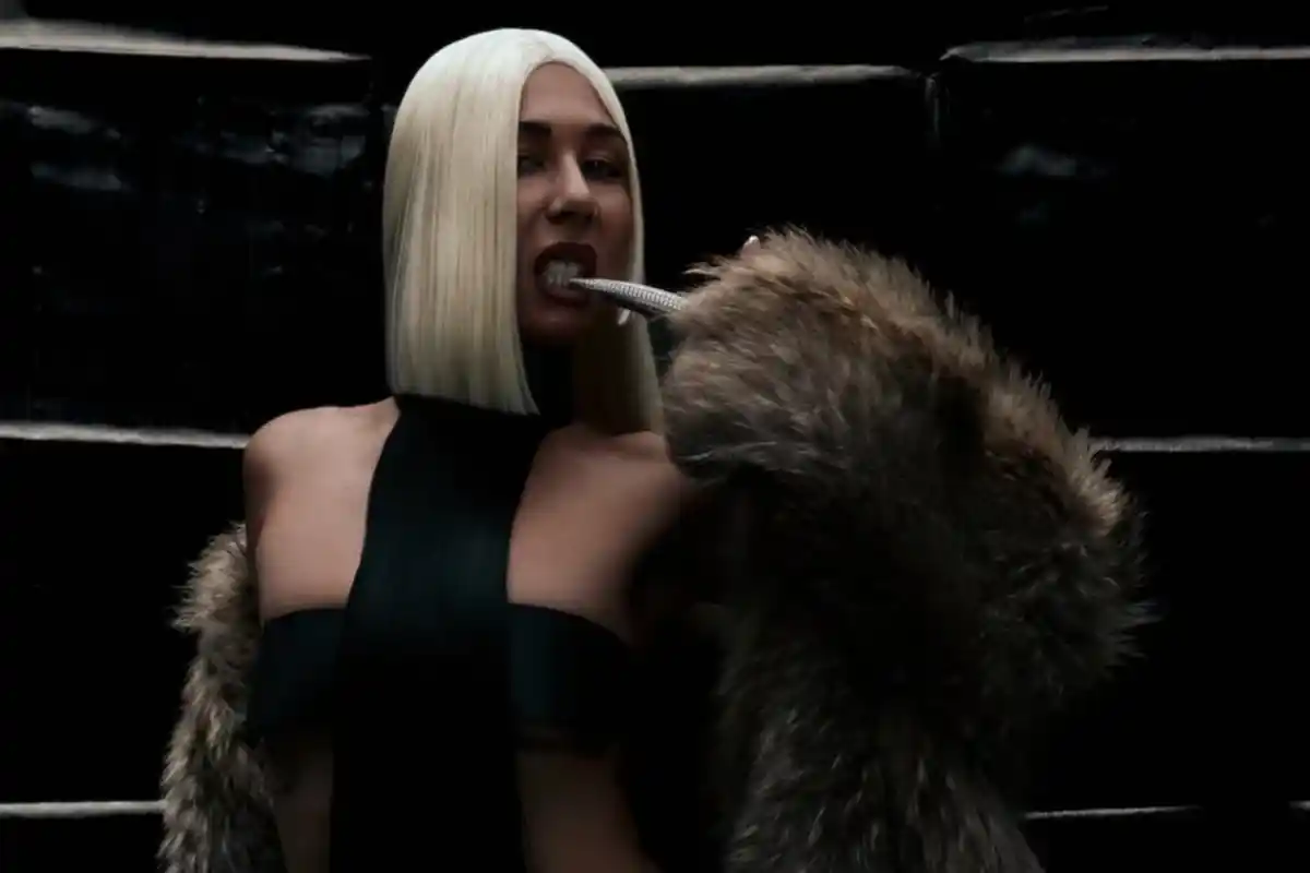 A dimly lit woman with a platinum blonde bob bites her crystal covered acrylic nail in while in a fur coat in front of a black wall.