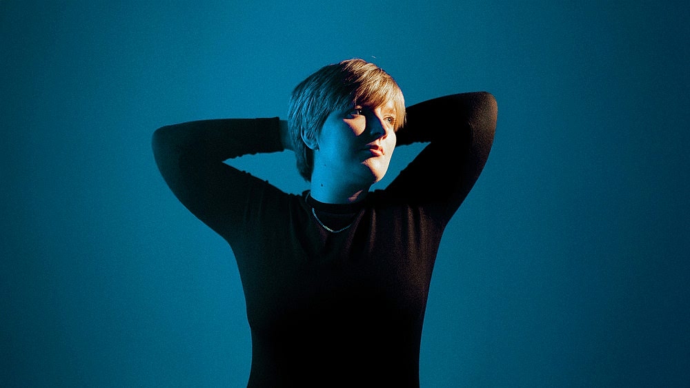 A person in a dark, long-sleeve shirt stands with arms raised behind their head, against a blue background